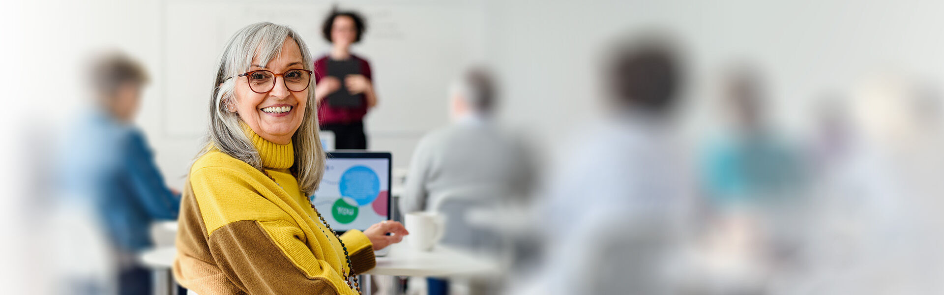Frau mit langen grauen Haaren sitzt in einem Schulungsraum und nimmt am Laptop an einem Coaching für über 60-Jährige teil.