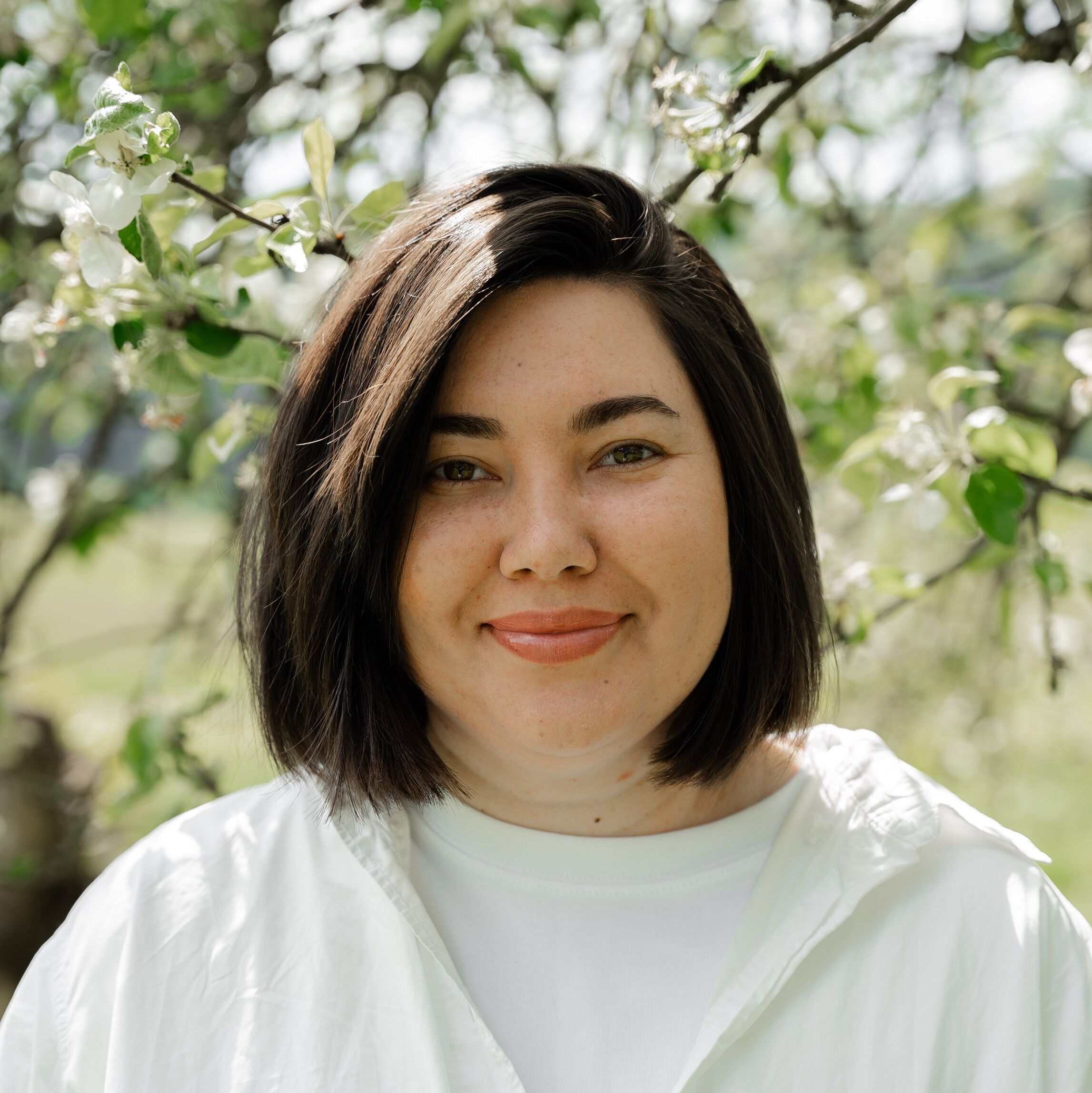 Foto von IBB-Dozentin Anzhelika Zavhorodnia, auf dem sie draußen in der Sonne vor einem blühenden Obstbaum steht.